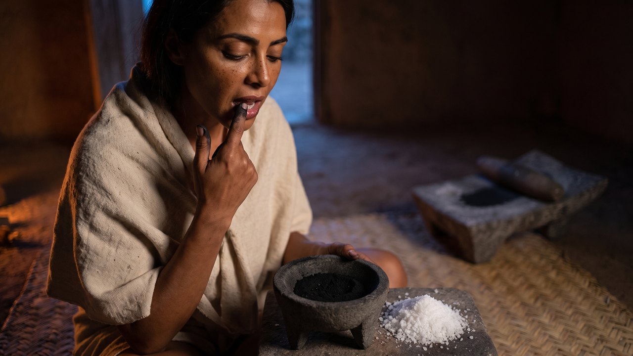 Is charcoal toothpaste good for your teeth — a Mexica woman in a house in Tenochtitlan presses a charcoal-blackened fingertip along her own gum, a three-legged basalt bowl of black charcoal dust and a heap of white salt beside her
